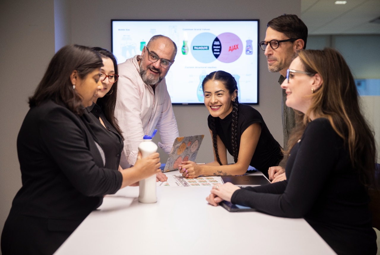 Un equipo trabajando en equipo. Al fondo, un panel interactivo con los logotipos de Palmolive y Ajax